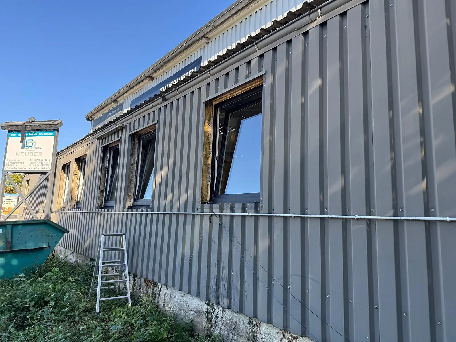 Reihe neuer anthrazitfarbener Fenster in der grauen Trapezblechfassade einer Halle, ein Flügel steht gekippt offen, die Laibungen sind noch offen