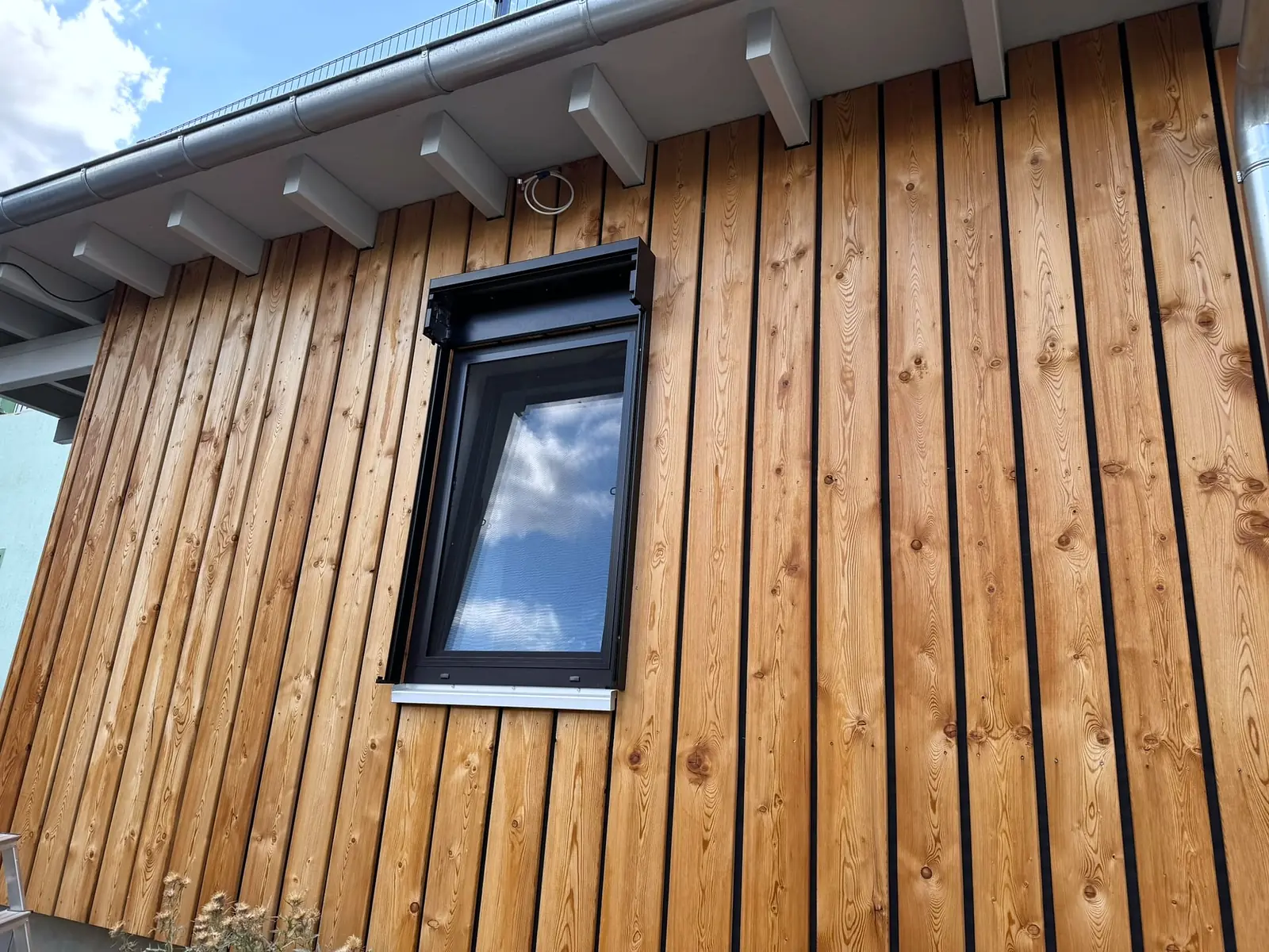 Gekipptes Fenster mit anthrazitfarbenem Rahmen und schwarzem Aufsatzkasten in einer Fassade aus senkrechten Lärchenbrettern