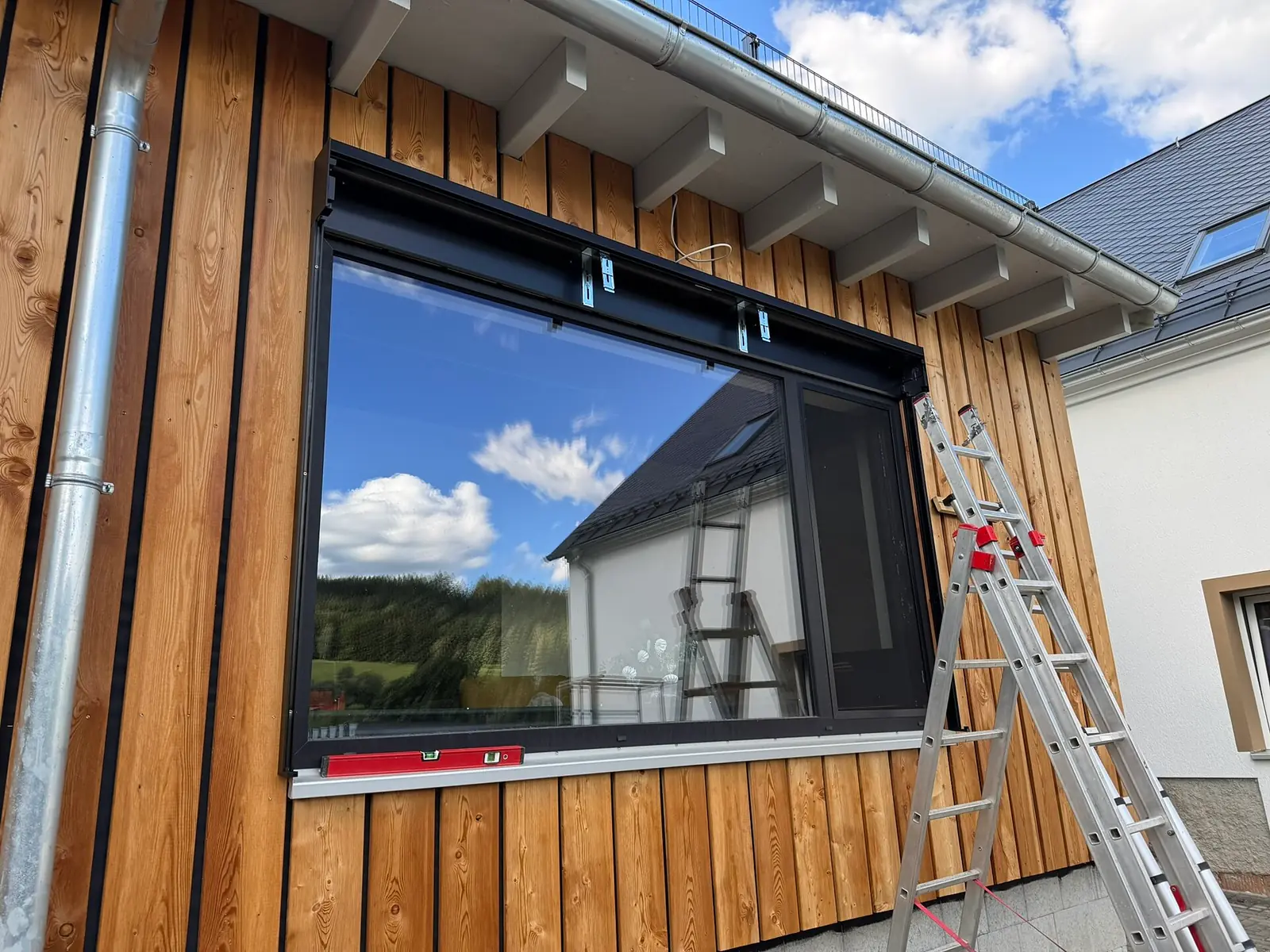 Breites Fensterelement mit anthrazitfarbenem Rahmen in einer senkrecht verschalten Holzfassade, davor eine angelehnte Aluleiter, auf der Fensterbank liegt eine Wasserwaage