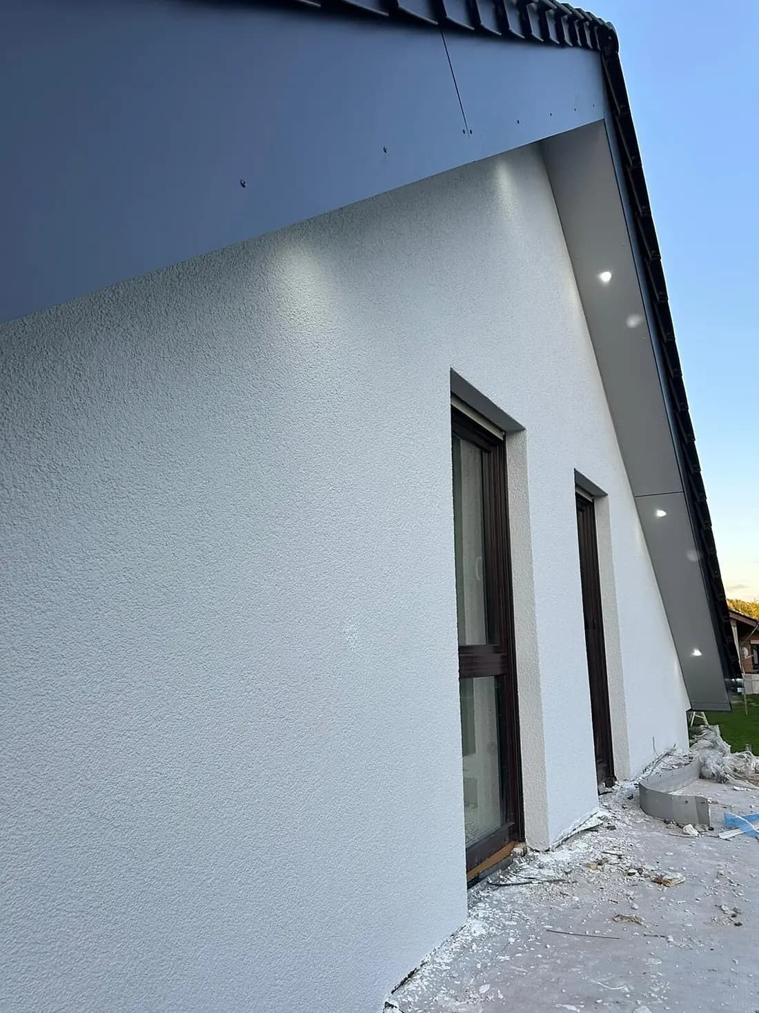 Weiß verputzte Hauswand unter einem dunkelgrauen Dachüberstand mit zwei schmalen, raumhohen Fenstertüren in braunem Rahmen, davor liegen Putzreste