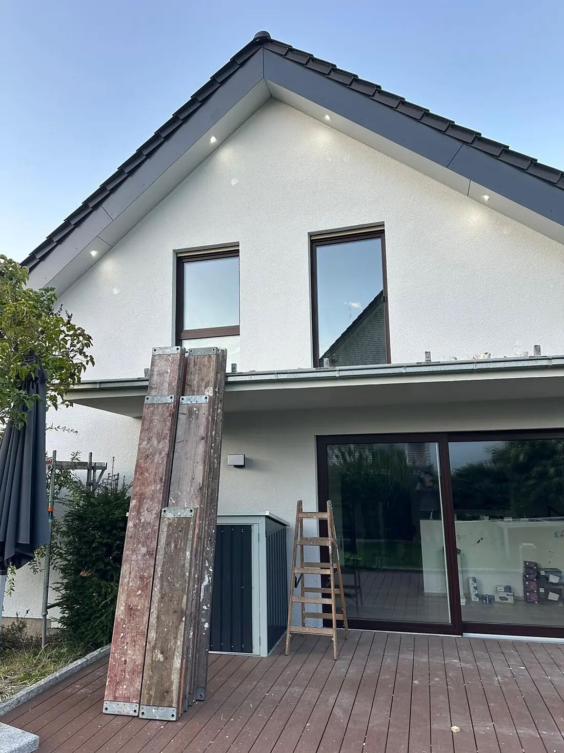 Giebelseite eines Wohnhauses mit weißer Putzfassade und dunkelgrauem Dach, oben zwei Fenster mit braunen Rahmen, unten eine große Hebeschiebetür zur Holzterrasse