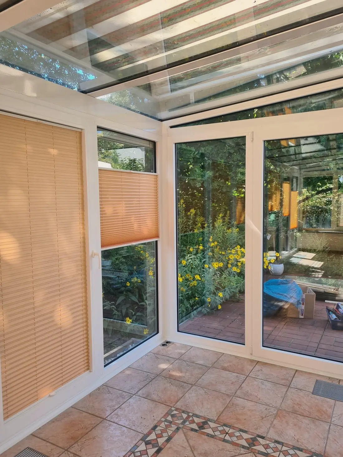 Ecke desselben Wintergartens von innen, durch die Fenstertüren sieht man in einen Garten mit gelb blühenden Stauden