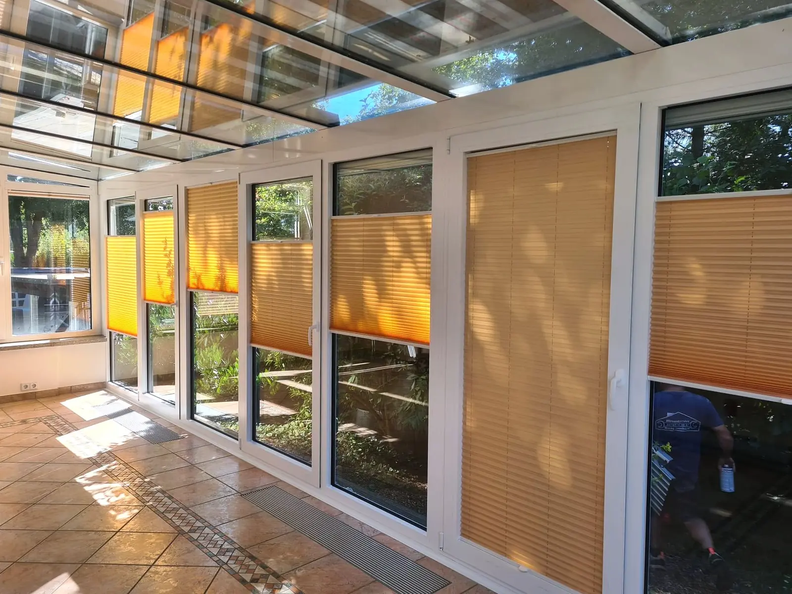 Wintergarten von innen mit Glasdach und einer Fensterfront, an der orangefarbene Plissees unterschiedlich weit zugezogen sind