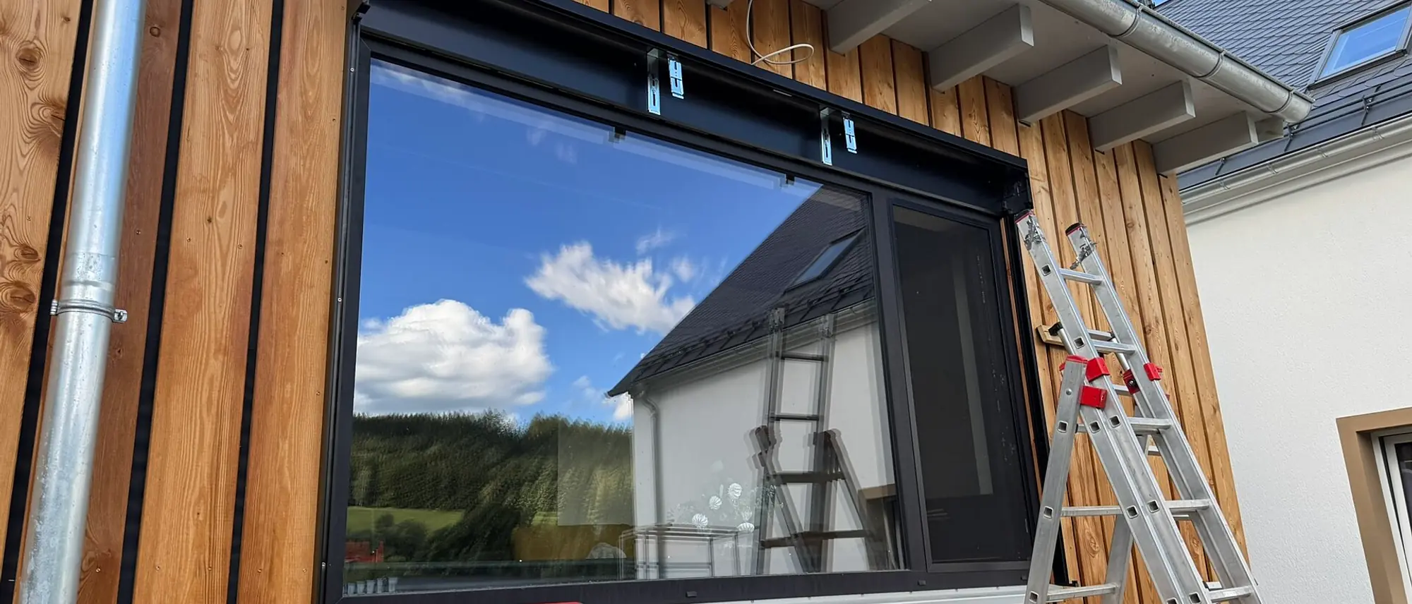 Breites Fensterelement mit anthrazitfarbenem Rahmen in einer Holzfassade, davor eine angelehnte Aluleiter